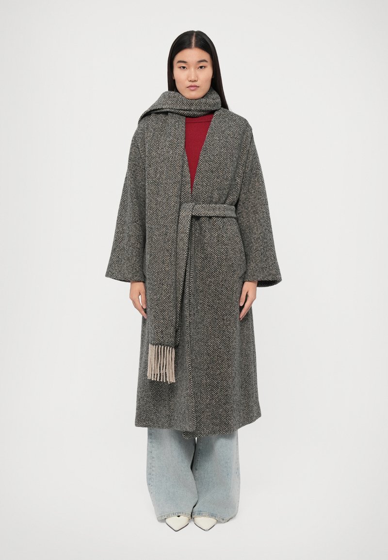 Manteau long en chevrons gris avec des manches larges et une écharpe assortie à bords frangés, superposé sur un top rouge et un jean bleu clair.