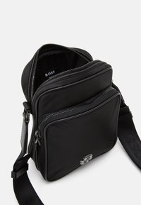 Sac bandoulière noir en tissu lisse, avec des compartiments zippés et un accent logo. Il dispose d'une sangle ajustable et d'un design rectangulaire élégant.