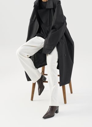 Schwarzer, oversized Trenchcoat über einem schwarzen Top, kombiniert mit weißen Hosen und braunen, spitzen Ankle Boots, auf einem Holzschemel sitzend.
