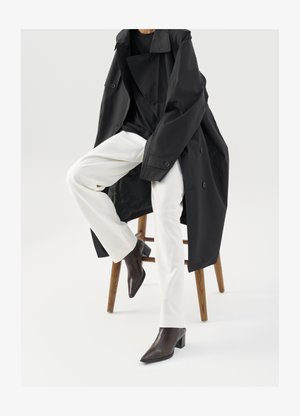 Abrigo trench negro y oversized sobre una camiseta negra, combinado con pantalones blancos y botines de punta marrones, sentado en un taburete de madera.