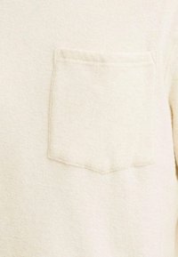 Maglione beige chiaro con un tessuto testurizzato, una tasca applicata sul lato sinistro del petto e cuciture visibili attorno ai bordi della tasca.