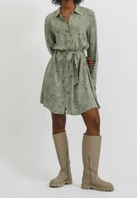 Kvinna klädd i en grön, prickig långärmad klänning med bälte och beige knähöga stövlar, som står mot en enkel ljus bakgrund.