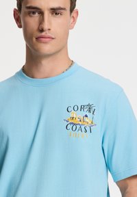 Svetlo modra bombažna majica z barvitim grafikami plažnih koč in palmami ter napisom "Coral Coast Shiwi" na prsih.