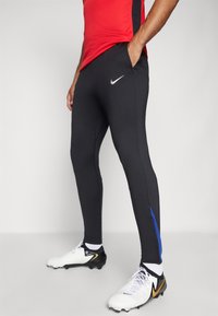 Czarne sportowe spodnie o dopasowanym kroju, z niebieskim paskiem po boku i srebrnym logo Nike z przodu. Wykonane z gładkiego, elastycznego materiału.