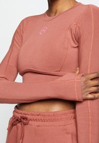 Yksityiskohta henkilöstä, joka on pukeutunut pölyisen vaaleanpunaisiin pitkähihasiin cropped-paitaan, jossa on peukalohourut, sekä yhteensopiviin naruhaalareihin.