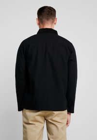 Czarna, długorękawowa kurtka o strukturalnym kroju, z kołnierzem, gładkim materiałem i jednolitymi plecami. W połączeniu z beżowymi spodniami.