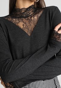 Woman wearing dark gray long-sleeve top with lace V-neckline and lace cuffs, paired with black-and-white houndstooth pants, arms crossed.