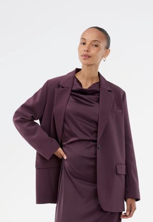 Overdimensional mørkeblå blazer med notched lapels og en enkelt knaplukning, båret over en matchende draperet satin-kjole. Glat tekstur.