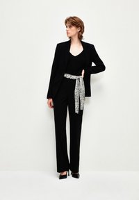 Blazer nero su un top nero, abbinato a pantaloni neri. Una cintura con stampa leopardata aggiunge contrasto. La modella indossa tacchi alti neri.