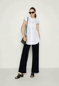 Camisa branca de botão com mangas curtas, cinto na cintura. Combinada com calças largas pretas e mules de salto aberto pretos.