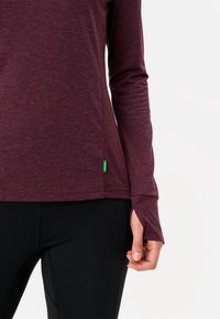 Maglietta a maniche lunghe in tessuto bordeaux con una texture melange, con un'etichetta logo verde sull'orlo e polsini con foro per il pollice. Indossata con leggings neri.