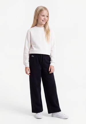 Fille souriante aux longs cheveux blonds, portant un sweat-shirt blanc, un pantalon noir à jambes larges et des baskets blanches, debout sur fond blanc.