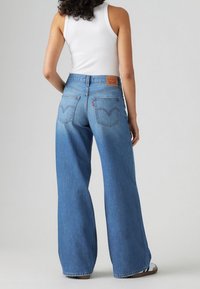 Kvinna som bär högmidjade vida blå jeans och en vit ärmlös topp, stående med ryggen mot betraktaren mot en enfärgad bakgrund.