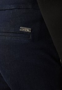 Gros plan de la poche arrière d’un jean en denim bleu foncé avec une petite plaque métallique rectangulaire comportant un emblème ailé gravé.