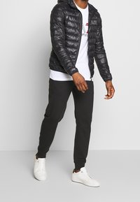 Svart pufferjacka med huva, vadderad design och dragkedjestängning, kombinerad med svarta joggingbyxor och vita sneakers. Vit t-shirt under.