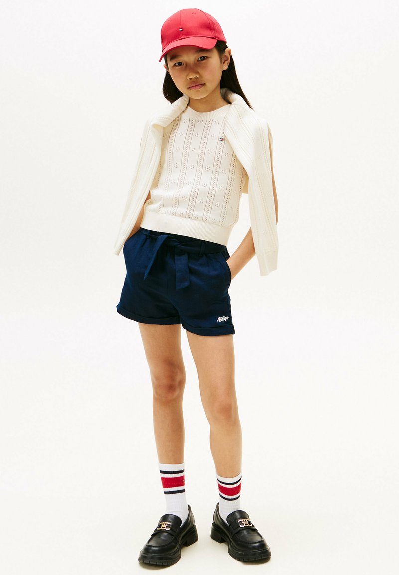 Débardeur tricoté blanc sans manches, shorts marine avec ceinture, casquette rouge, mocassins noirs et chaussettes rayées. Design simple et esthétique décontractée.