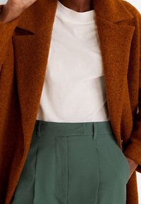 Verbrand oranje wollen jas met een textuurafwerking, gelaagd over een wit T-shirt, gecombineerd met op maat gemaakte groene broeken met plooien.