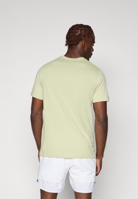 Svetlozelen majica iz bombaža s kratkimi rokavi in okroglim izrezom, ki ima gladko teksturo, prikazana s hrbtne strani, v kombinaciji z belimi kratkimi hlačami.