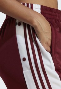 Pantalones deportivos granate con rayas laterales blancas, que cuentan con acentos de botones y un bolsillo frontal. Están hechos de una tela suave con un leve brillo.