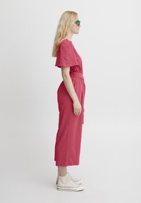 Tuta rosa con maniche corte, vita con cintura e gambe larghe, abbinata a sneakers bianche e occhiali da sole, presentata su uno sfondo neutro.