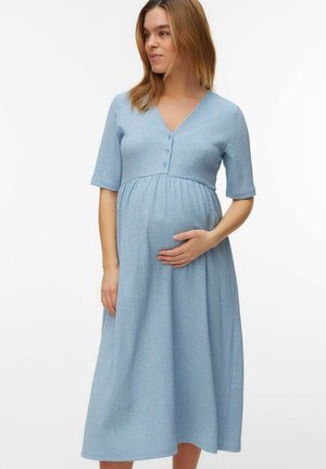 Kobieta w ciąży ubrana w jasnoniebieską, teksturowaną sukienkę midi z krótkimi rękawami, stojąca i delikatnie trzymająca brzuch.