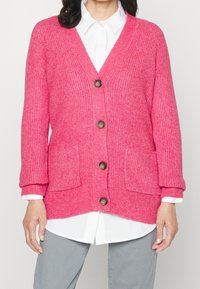 Rosa kofta med ribbad textur, knappframsida och två framfickor. Har avslappnad passform och uppvikta ärmar över en vit skjorta.