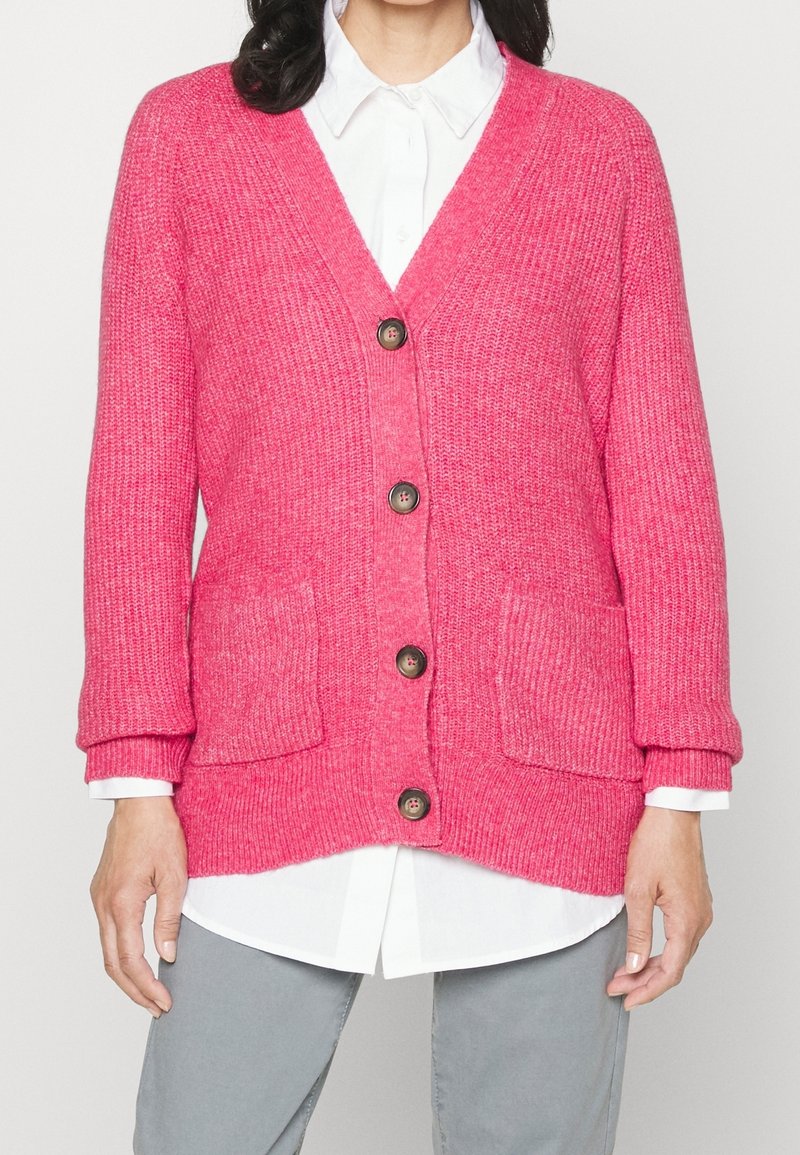 Rosa kofta med ribbad textur, knappframsida och två framfickor. Har avslappnad passform och uppvikta ärmar över en vit skjorta.