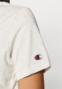 Κοντομάνικο, ανοιχτό μπεζ μπλουζάκι από μαλακό ύφασμα. Το μανίκι διαθέτει ένα μικρό κεντημένο λογότυπο με κόκκινες και μαύρες λεπτομέρειες.