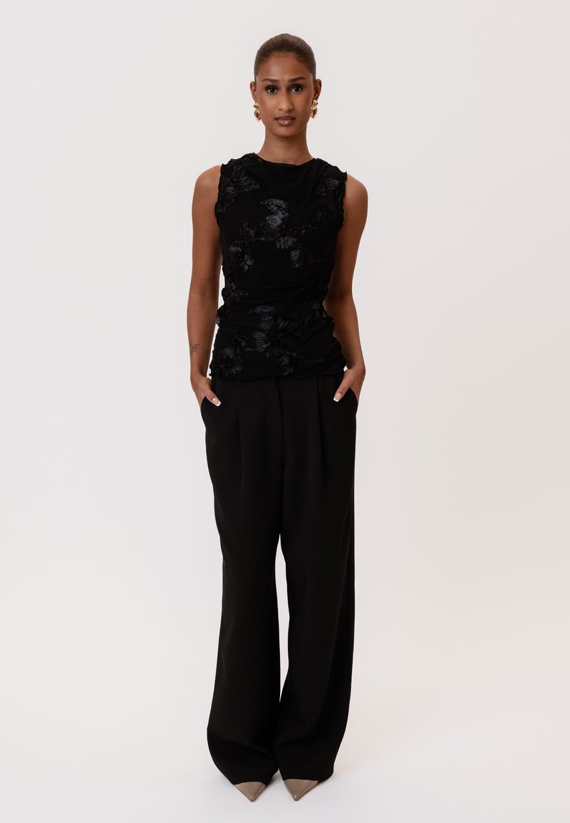 Femme debout les mains dans les poches, portant un haut noir sans manches avec des détails floraux et un pantalon noir à jambes larges sur fond blanc.
