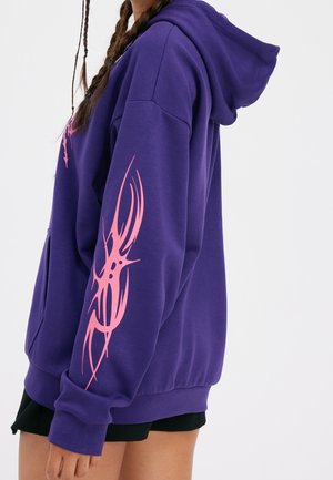 Personne portant un sweat à capuche violet avec des motifs tribaux roses sur la manche et un short noir sur un fond blanc uni.