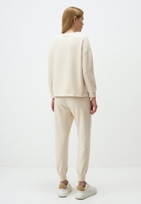 Felpa beige con spalle scese, maniche con polsini e pantaloni a sigaretta coordinati. Texture liscia, design minimal e polsini elastici sui pantaloni.
