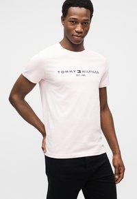 Ανοιχτό ροζ βαμβακερό T-shirt με λογότυπο σε ναυτικό μπλε με κόκκινες αποχρώσεις, στρογγυλή λαιμόκοψη, κοντά μανίκια και εφαρμοστή σχεδίαση. Φορεμένο με μαύρο παντελόνι.