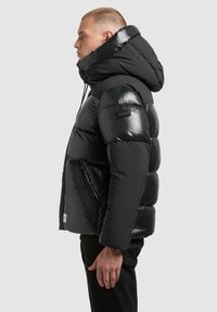 Chaqueta acolchada negra con acabado brillante, capucha grande, bolsillos con cremallera y parche de logo rectangular en el hombro izquierdo.