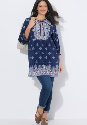 Mujer sonriente con cabello largo y oscuro, vistiendo una túnica azul con motivos, jeans azules, zapatos marrones y llevando un bolso beige.