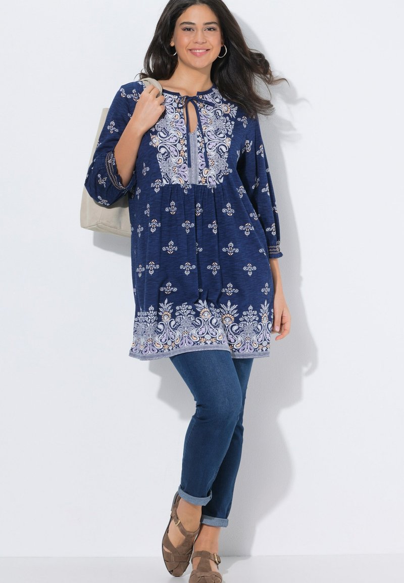 Lächelnde Frau mit langen dunklen Haaren, die eine blau gemusterte Tunika, blaue Jeans, braune Schuhe trägt und eine beige Handtasche hält.