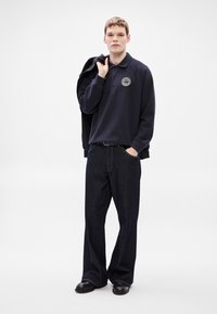 Μπλε σκούρο πόλο πουκάμισο με λογότυπο, συνδυασμένο με σκούρο, φαρδύ τζιν παντελόνι. Το μοντέλο κρατάει ένα μαύρο σακάκι πάνω στον έναν ώμο, φοράει μαύρα παπούτσια.