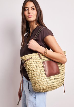 Donna con indosso un top scuro e jeans chiari che porta una grande borsa tote intrecciata in paglia con dettagli in pelle marrone sulla spalla.