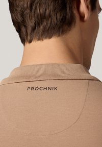 Beżowa koszulka polo z teksturowanym wzorem dzianiny, z prążkowanym kołnierzykiem i czarnym logo "PROCHNIK" wyszytym na plecach.