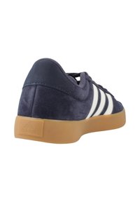 Marineblauwe suède sneaker met gumrubberen zool, witte zijstrepen, veters en een gewatteerde kraag, gezien vanuit een achter-linkerhoek.