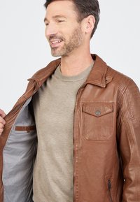 Braune Lederjacke mit einem Reißverschluss vorne, zwei aufgesetzten Taschen und einem grauen Innenfutter. Darunter ein hellbeiges Sweatshirt. Glatte Textur und strukturierter Schnitt.