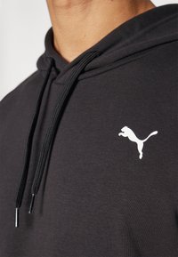 Nærbillede af en person iført en sort hættetrøje med sorte snore og et hvidt Puma-logo på brystet.