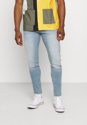 Mann trägt hellblaue Skinny-Jeans, weiße High-Top-Sneaker und ein kurzärmeliges Hemd mit Knöpfen in Gelb-, Schwarz-, Weiß- und Olivtönen.