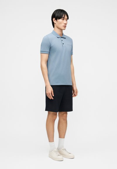 Άνδρας όρθιος, φορώντας ανοιχτό γαλάζιο πόλο πουκάμισο, μαύρο σορτς, άσπρες κάλτσες και άσπρα αθλητικά παπούτσια, με φόντο ένα απλό ανοιχτό χρώμα.