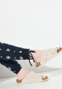 Jasnobeżowe zamszowe klapki z dwoma regulowanymi paskami z klamrami, korkowa wkładka i biała gumowa podeszwa, noszone z granatowymi legginsami w wzory.