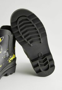 Bottes noires imperméables avec une semelle en caoutchouc texturée arborant un motif en zigzag et des accents jaunes éclaboussés sur le côté.