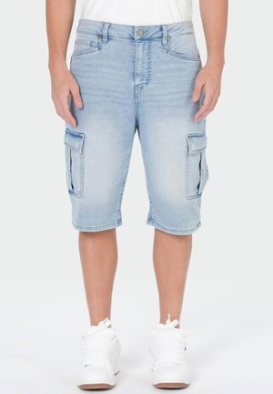 Short cargo en denim bleu clair avec poches à rabat latérales porté par une personne debout, associé à des baskets blanches et un haut blanc.