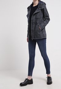 Navy-mönstrad jacka med huva, metalldragkedja framtill och sidofickor; kombinerad med mörka jeans och svarta skor. Slätt tyg med geometrisk design.