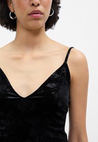 Melnas samta topiņš ar ziedu tekstūru, dziļu V-veida kakla izgriezumu un plānām regulējamām siksnām. Iezīmē cieši pieguļošu dizainu.