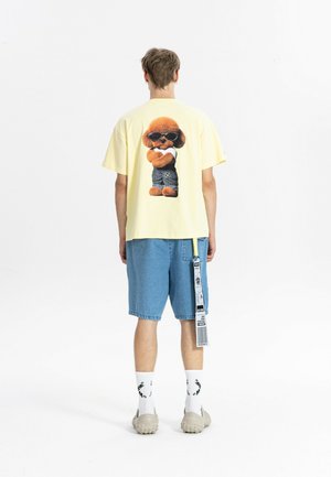 Hombre que lleva una camiseta amarilla con un gráfico de un perro con gafas de sol en la espalda, combinado con pantalones cortos vaqueros azules y calcetines blancos.