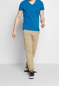 Blaues V-Ausschnitt-T-Shirt kombiniert mit beigen Chinos. Das Modell trägt schwarze Sneakers, hat kurze Ärmel und ein sauberes Design ohne sichtbare Muster.